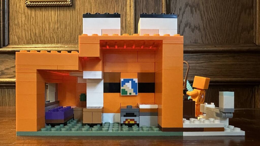 This is an image of the rear view of the Fox Lodge. There is an open back and on the interior is a purple bed, with a plant beside it. There is a crafting table, a furnace, a cake and a map. This is from the LEGO Minecraft Fox Lodge Set
