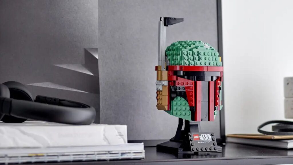 The Boba Fett helmet is on a desk, next to. a pile of books and headphones.